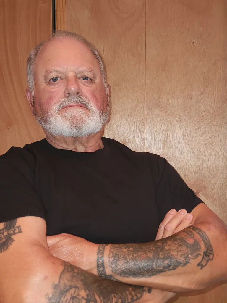 Portrait of Frank, a personal trainer with over 25 years of experience, wearing a black t-shirt with arms crossed, displaying professional confidence and expertise.